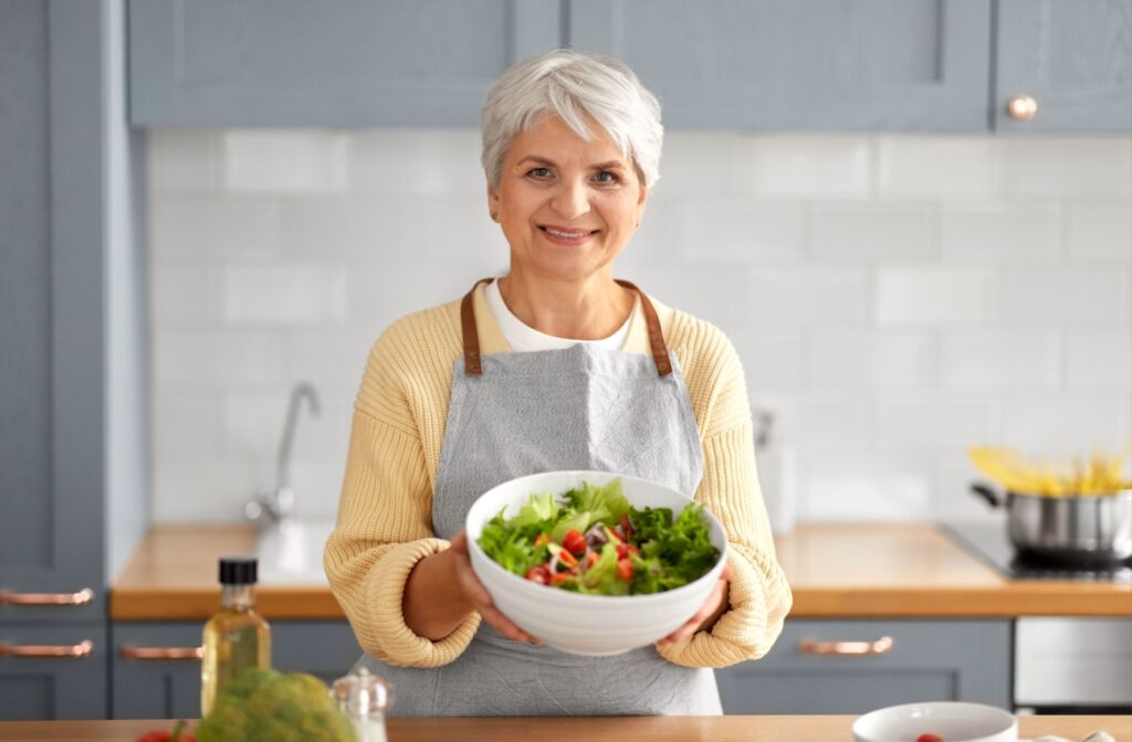 An older adult woman in a kitchen holding a bowl of salad with both hands, smiling and looking directly at the camera.