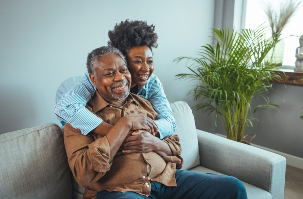 Older adult sitting on a couch as his daughter hugs him, both smiling.