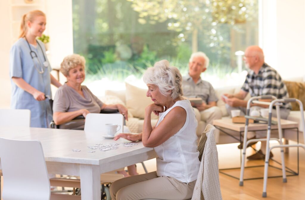 An older adult woman sitting at a table doing a puzzle in a senior living community with other residents in the background.